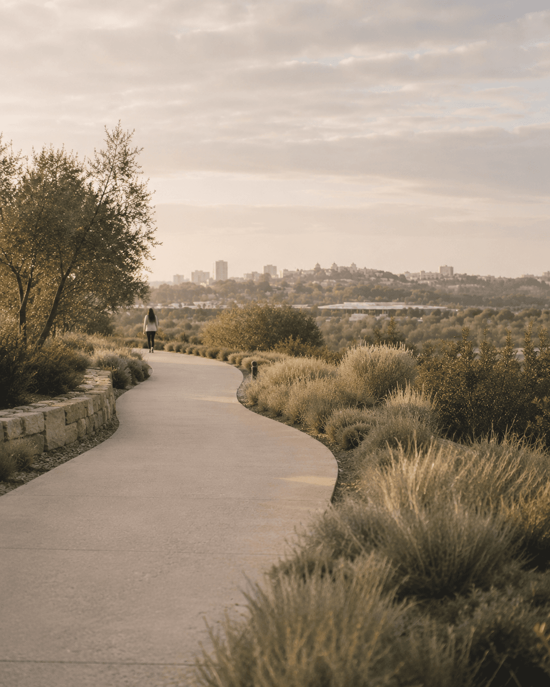 A person walking along a winding path through native grasses with a city skyline in the distance at golden hour